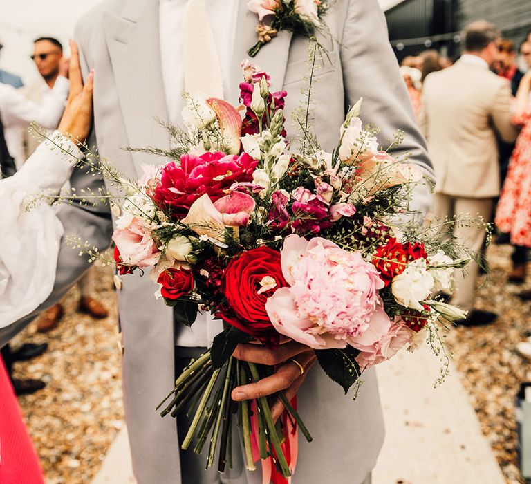 groom in a light grey suit holding a pink peony an red rose wedding bouquet at coastal wedding 