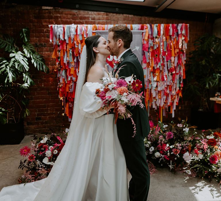 Bride in Suzanne Neville wedding dress with detachable sleeves kissing groom in green suit with rainbow streamers 