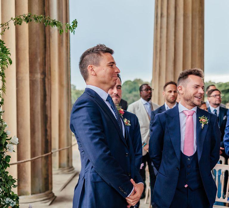 Groom in blue suit smiles as he sees the bride walking down the aisle 