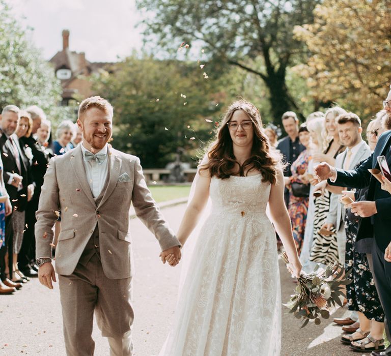 Groom in grey wedding suit and green bow tie walks hand in hand with the bride for their confetti exit moment 