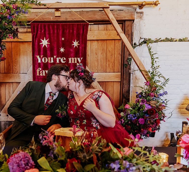 Dark red and gold wedding banner sign backdrop to the sweetheart table with the bride and groom sharing a kiss 