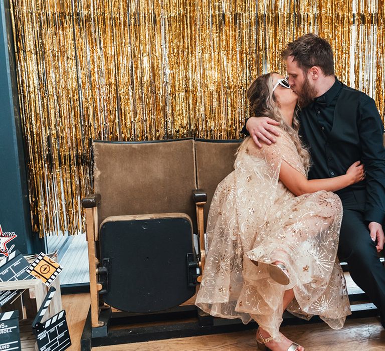 Bride in gold metallic wedding dress with groom in all black wedding suit kissing as they pose for the photobooth 