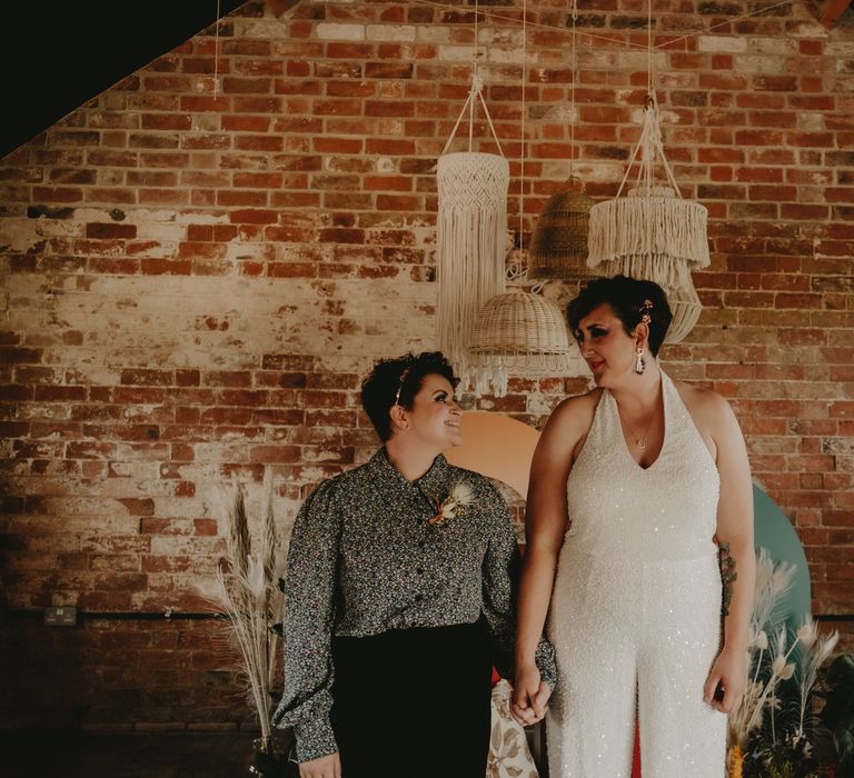 Bride in a jumpsuit and bride in a patterned shirt and trousers holding hands 