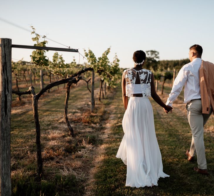 Bride and groom walking through a Vineyard in the UK holding hands. The bride wears separates and a white headdress