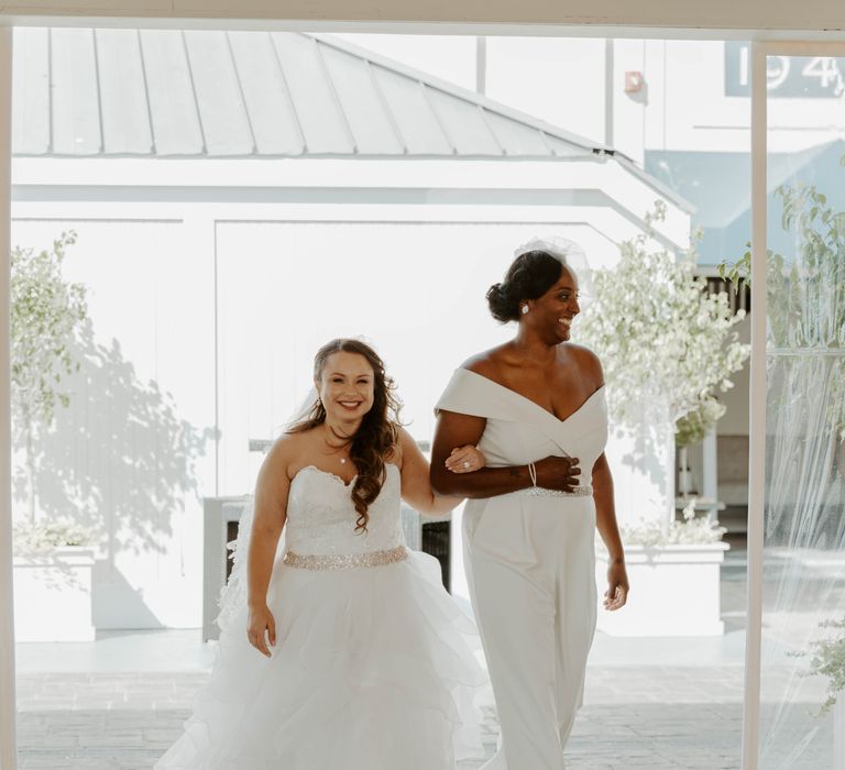 Black bride in a bridal jumpsuit and bi-racial bride in a strapless wedding dress entering the wedding reception as wife an wife 