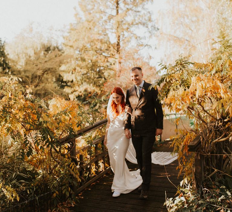 Bride and groom walking over wooden bridge surrounded by autumnal leaves. 