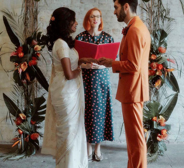 Celebrant lead wedding ceremony with bride in a white sari and groom in an orange suit exchanging their vows