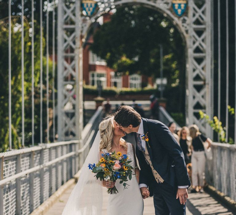 Bride in Justin Alexander wedding dress holding colourful bouquet kisses groom on bridge in Chester