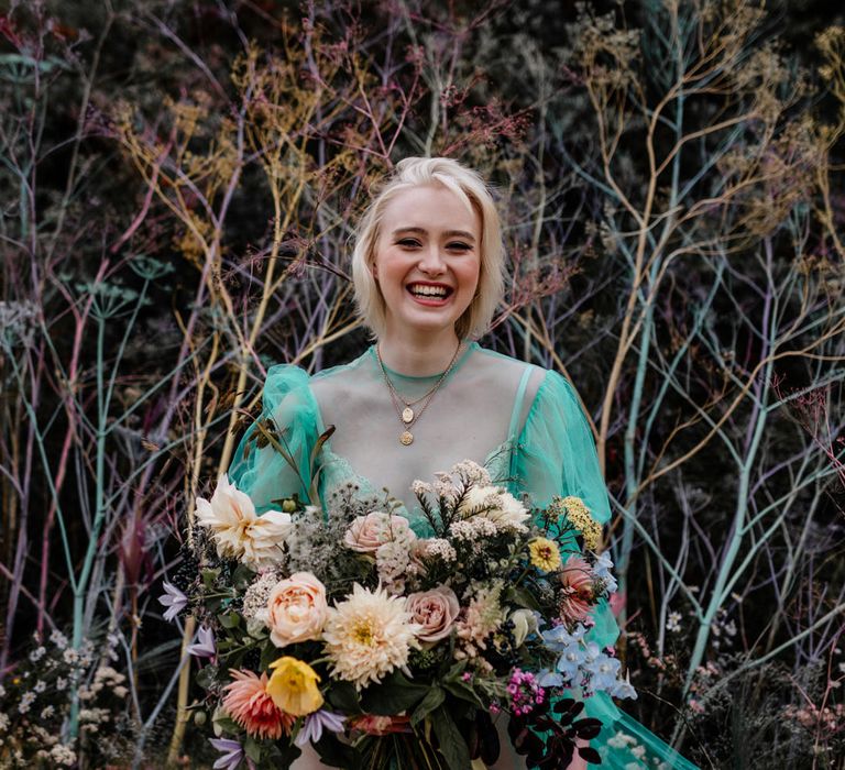 Smiling blonde bride in a high neck sheer wedding dress wearing gold chains and holding a colourful wedding bouquet 