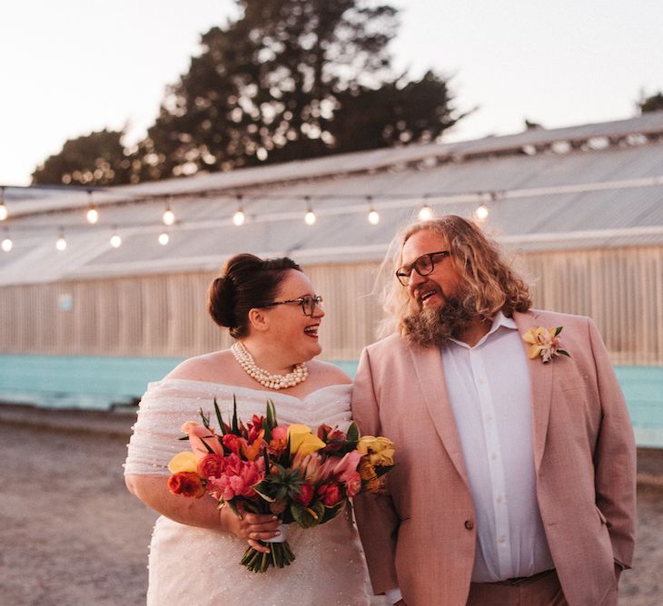 Bride in an off the shoulder wedding dress holding a colourful wedding bouquet looking at her bearded groom in a pink suit 