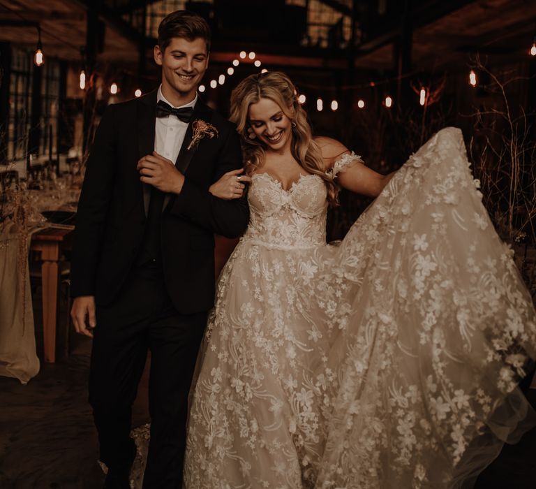 Bride in an intricate lace wedding dress holding out her skirt as she walks through the festoon lit reception with her groom 