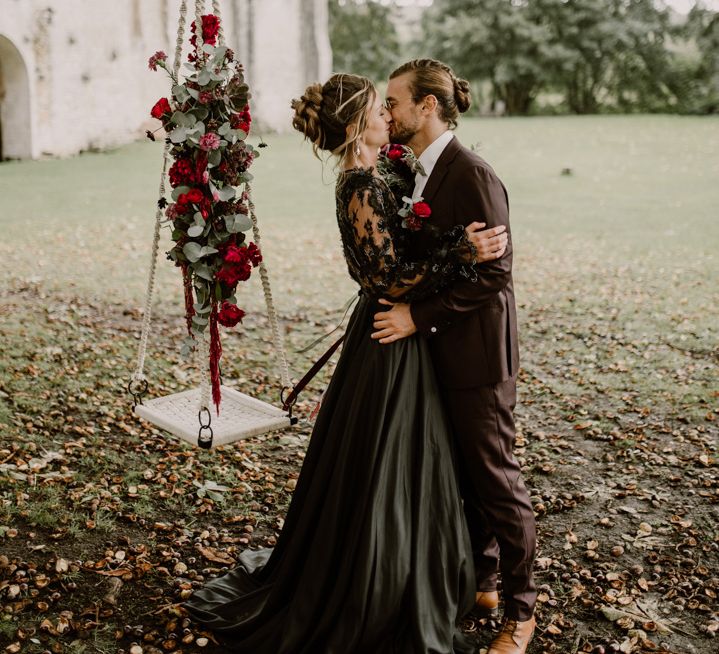 Alternative bride and groom in a black wedding dress and coloured suit kissing by a tree swing decorated with red flowers 