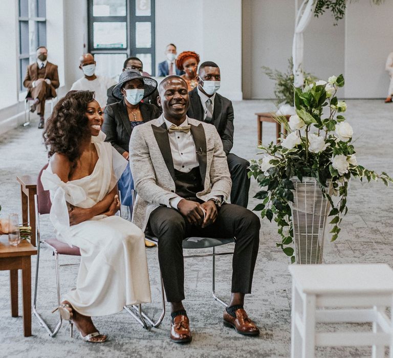 Smiling bride in white Bec + Bridge wedding dress sits next to smiling groom in grey suit jacket, black trousers and brown shoes at Bridge Community Church wedding