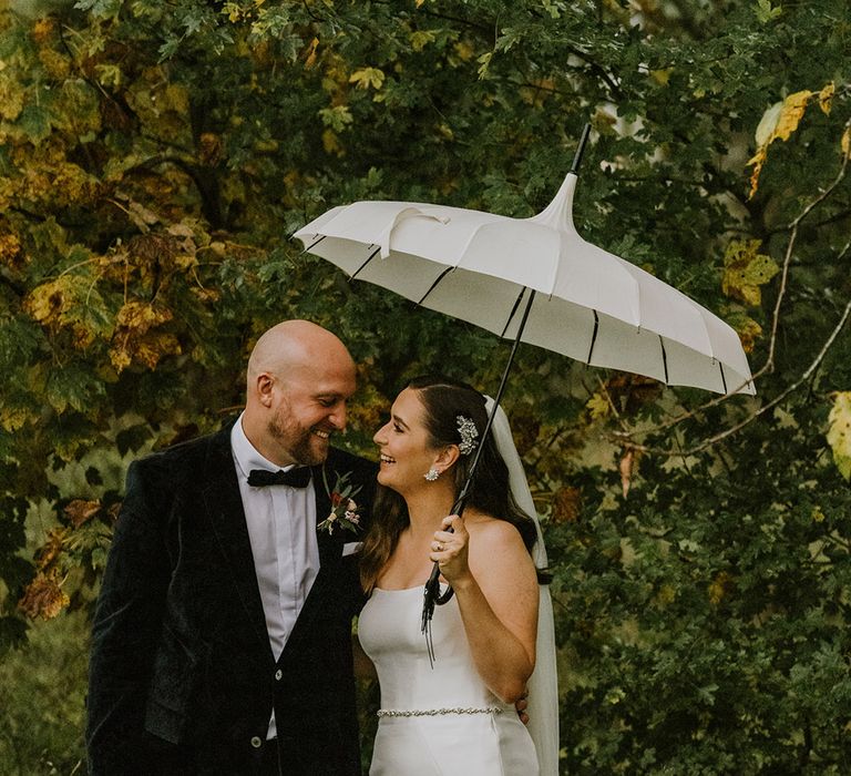 Portrait of the bride and groom in a velvet tuxedo and strapless wedding dress standing under an umbrella 