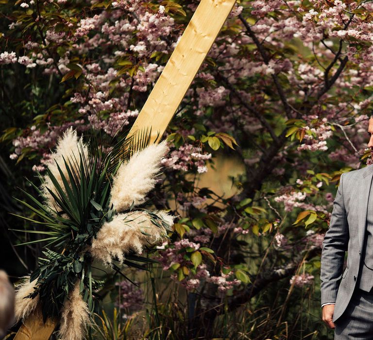 Groom in grey Hugo Boss suit with matching waistcoat and yellow tie with orange pocket square stands in front of wooden triangular wedding arch decorated with palm leaves and pampas grass