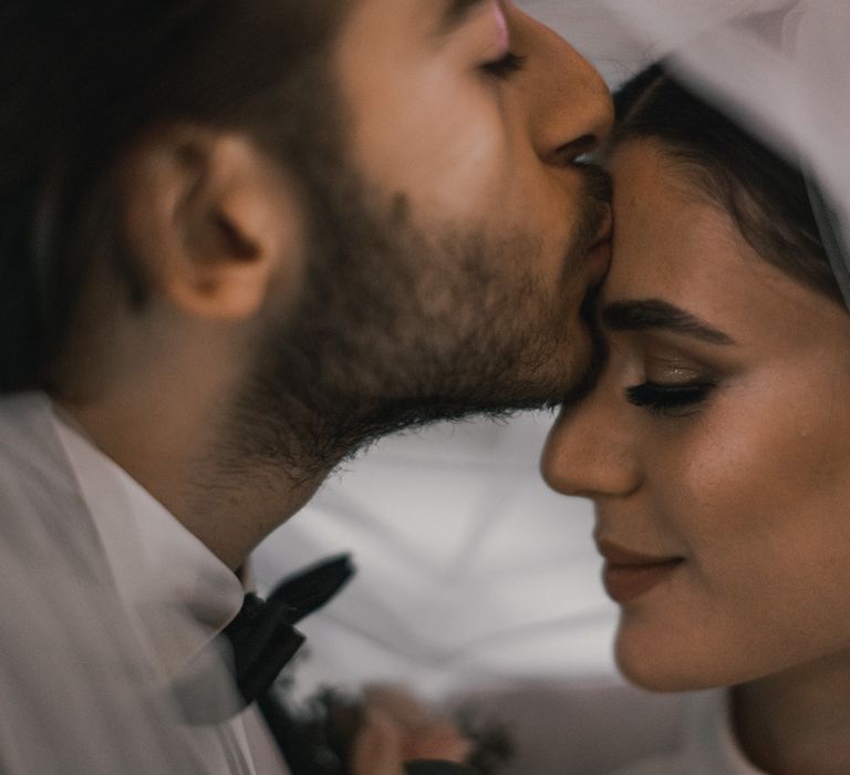 Groom kisses the forehead of his bride as they stand under her veil