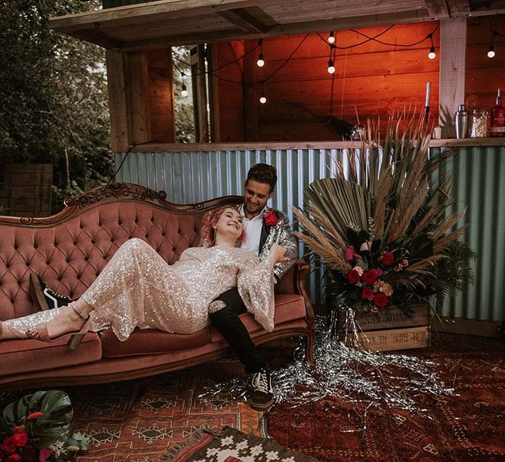 Bride in gold sequinned wedding dress lying on velvet sofa outside with groom in silver sequinned suit jacket at festival themed wedding