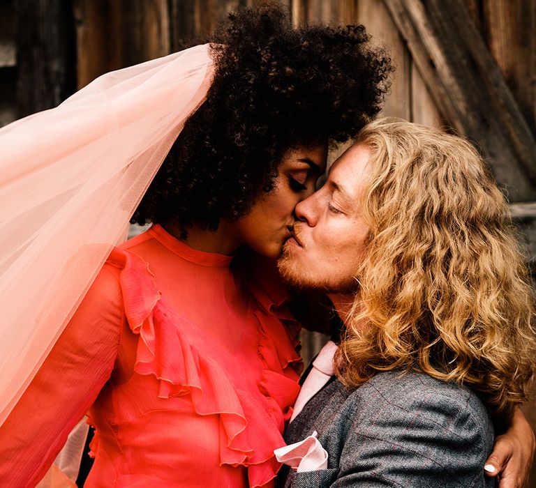 Bride in a red ruffle dress kissing her groom in a grey check suit on a motorbike 