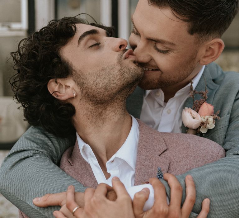 Grooms kiss outdoors whilst holding arms around one another