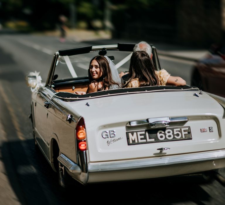 Bride in Rebecca Vallance Dress looks behind her as she rides in vintage Triumph convertible to Harrogate wedding