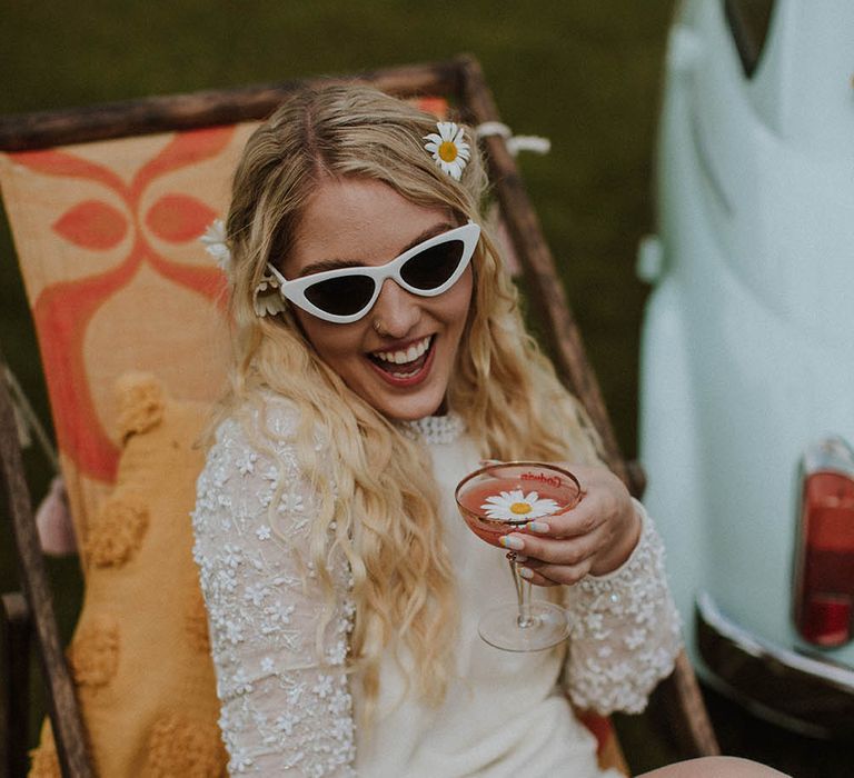 Bride in a short wedding dress with appliqué long sleeves holding a coupe glass and wearing white winged sunglasses 