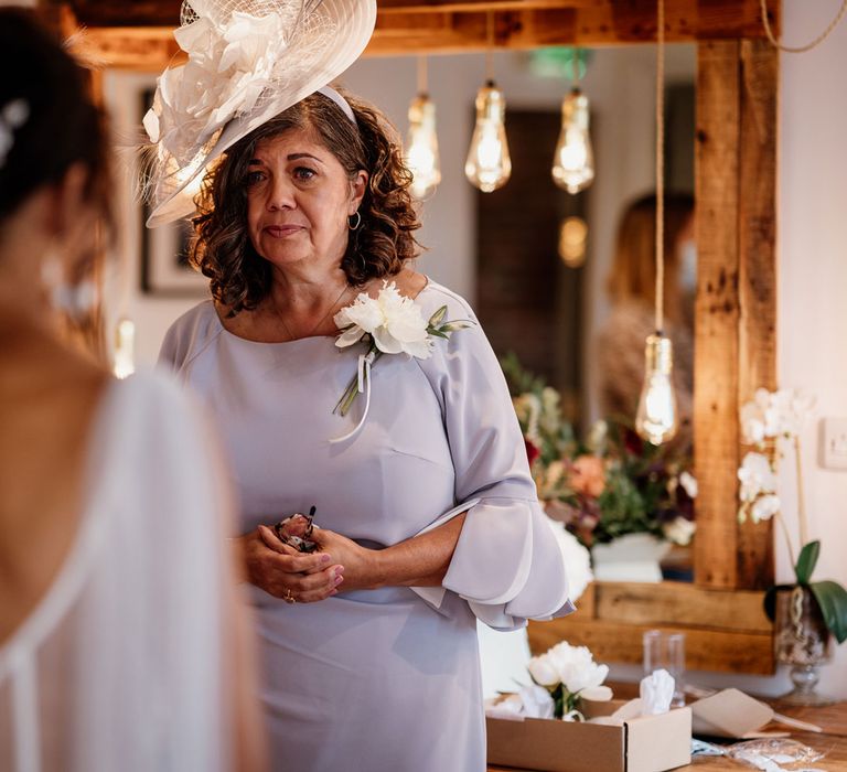 Emotional mother of the bride in light purple long sleeved dress, white corsage and white wedding hat looks at bride in low mesh back wedding dress before ceremony at The West Mill Derby