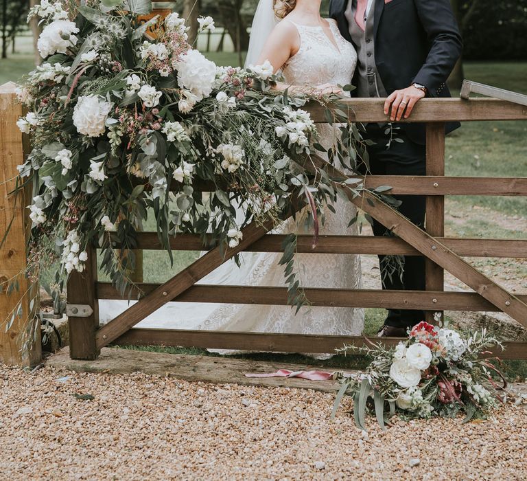 Bride in white lace Justin Alexander wedding dress kisses groom in dark three piece suit with grey waistcoat and pink tie behind wooden gate with white, pink and green floral drapery at Tythe Barn wedding with barn wedding flowers