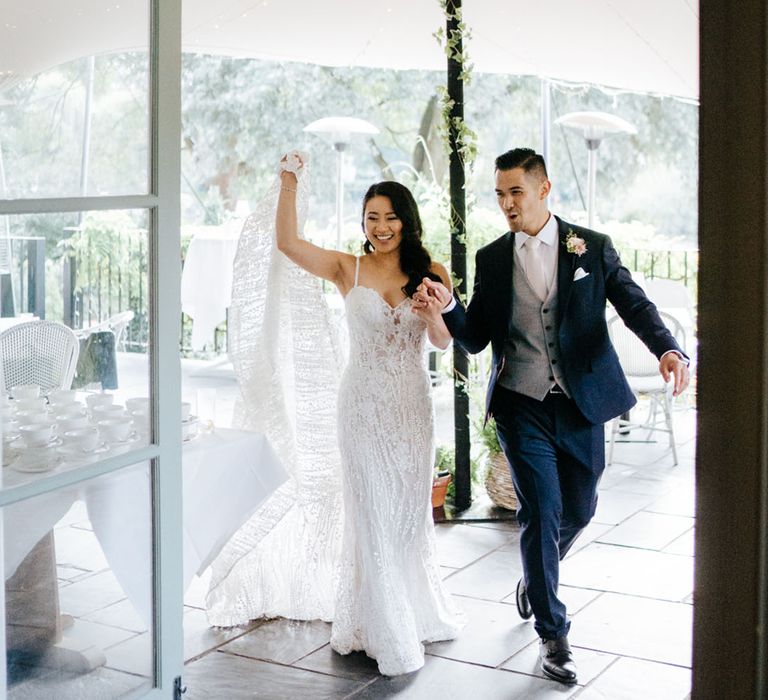 Bride in a mermaid wedding dress with thin straps holding hands with her groom in a navy suit as they walk into their Bingham Riverhouse Wedding