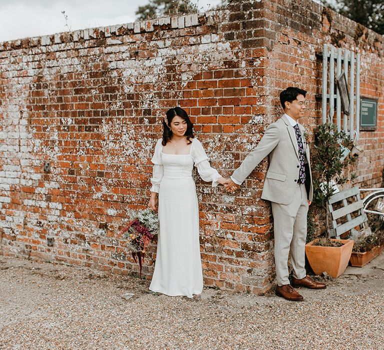 Bride & groom stand beside brick wall as they hold hands 