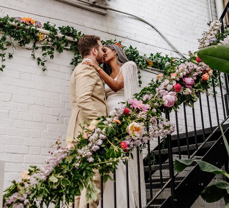 Bride in a feather Halfpenny London Cape kissing her groom in a beige suit on the stairs at The Loft Studios decorated in colourful flowers 