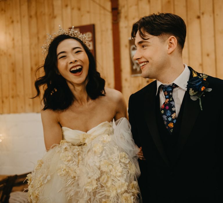 Bride in strapless wedding dress and bewjelled headpiece laughs with groom in blue suit, floral tie and floral buttonhole for rustic wedding