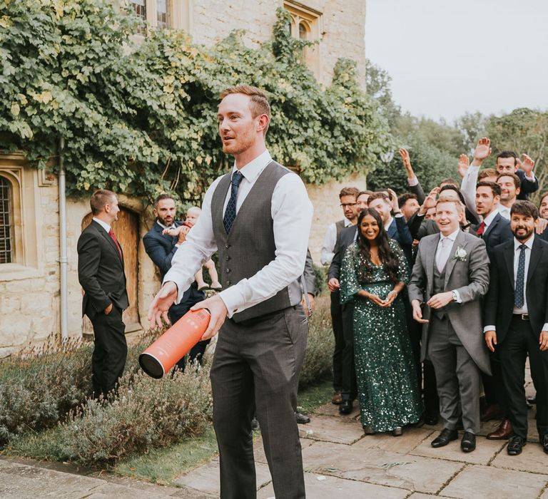 Groom in grey waistcoat and suit trousers gets ready to throw whisky bottle box to groomsmen, wedding guests and women in green sequin dress