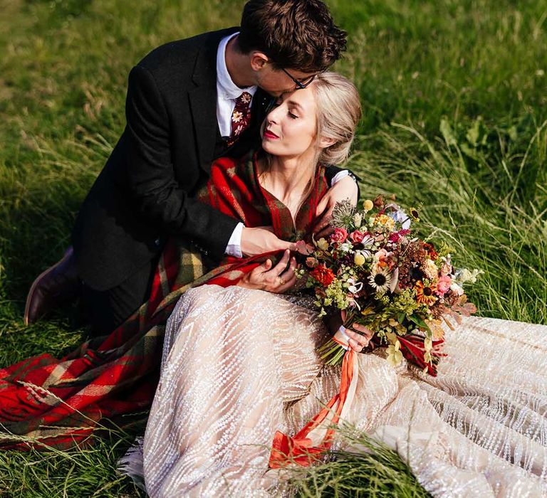 Bride & groom sit on the grass with one another as the groom wraps his arms around his bride