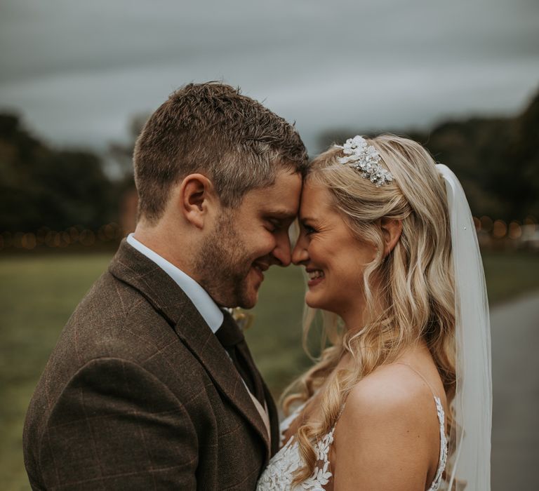 Bride in lace wedding dress and veils stand facing groom in brown check suit outside after barn wedding ceremony