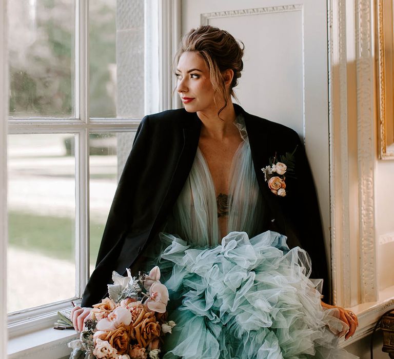 Beautiful bride with a pinned up do and natural makeup up sitting in a window frame in a light blue wedding dress and jacket holding a blush wedding bouquet