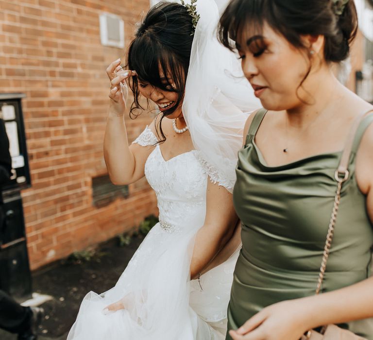 Bride in lace bardot wedding dress with pearl necklace and veil walks with bridesmaid in sage green satin bridesmaid dress towards wedding venue