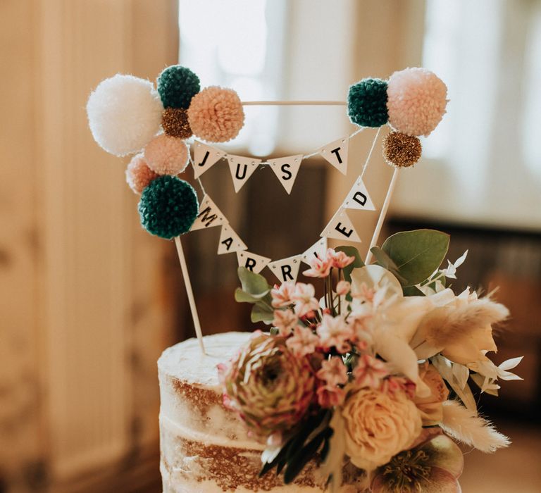 Rustic semi naked wedding cake with floral decor, bunting wedding topper reading 'Just Married' and pom poms