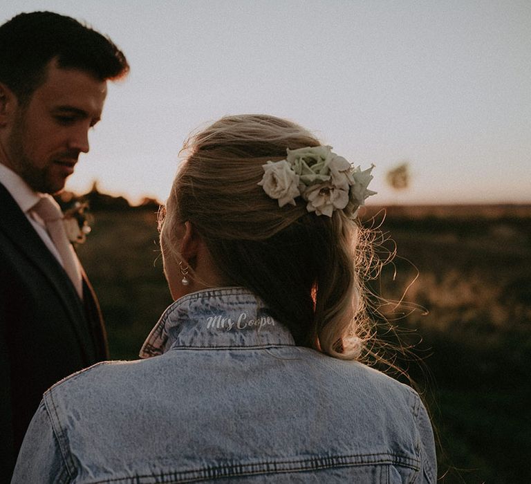 Bride in personalised denim jacket with 'Mrs Cooper' under collar of jacket and flowers in elegant half up half down hair with groom 