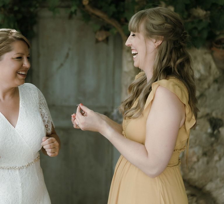 Bride with pearl earrings and lace jumpsuit stands with wedding guest in yellow floaty dress 