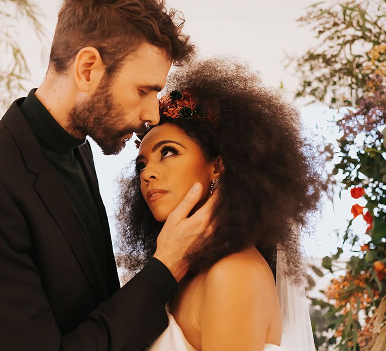 Groom in a black suit and top embracing his Black brides face with afro hair, off the shoulder wedding dress and winged eyeliner 