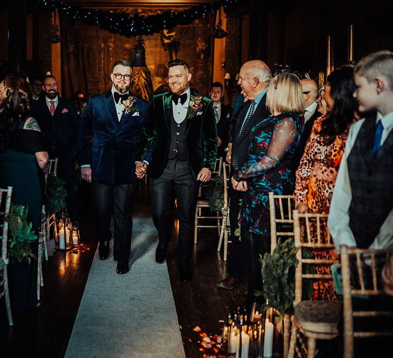 Grooms in velvet suit jackets and matching black bow ties walk the aisle hand in hand with candles and rose petal aisle decor