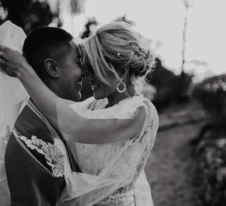 Bride in a lace wedding dress with pearl hoop earrings and pearl hair pins embracing her partner in a black tuxedo 