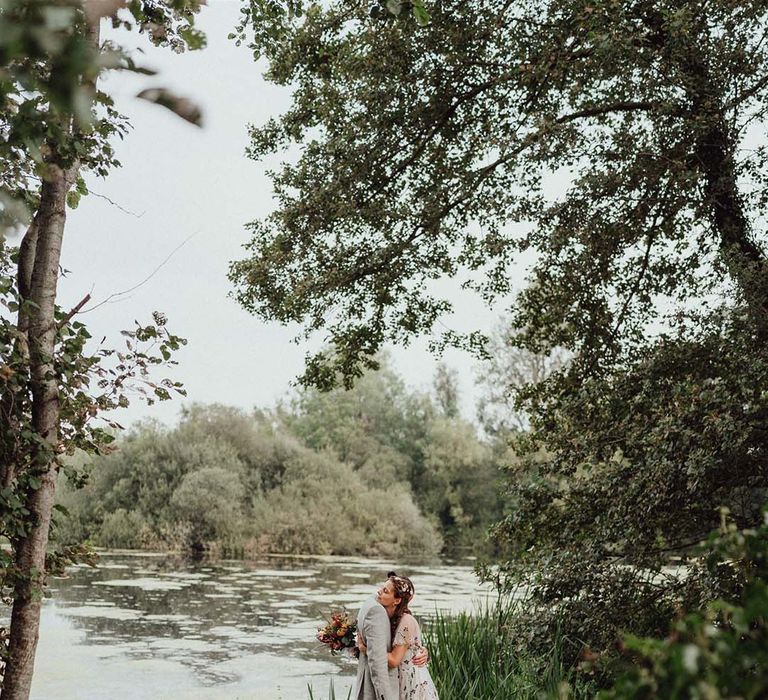 Bride in floral embroidered dress hugs groom in grey suit on jetty next to lake at wedding venue 