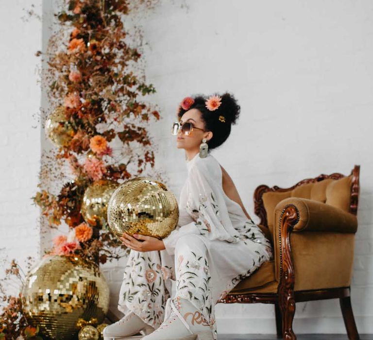 70s disco wedding inspiration with Black bride in a floral jumpsuit and roller skates sitting on an ornate couch holding a gold disco ball