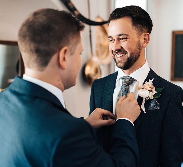 Groomsman helps fix the groom's paisley tie in dark blue suit with neutral flower buttonhole 