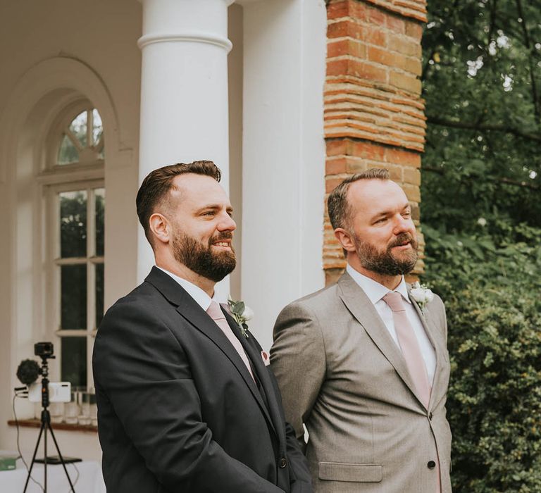 Groom in dark suit and groomsmen in light grey suit and pink ties get their first look at the bride