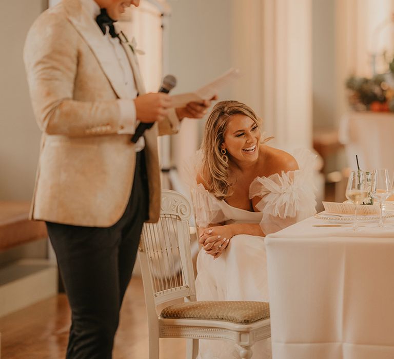 Bride in ruffle sleeve wedding dress laughs at the groom's speech who wears black tie and custom gold tuxedo