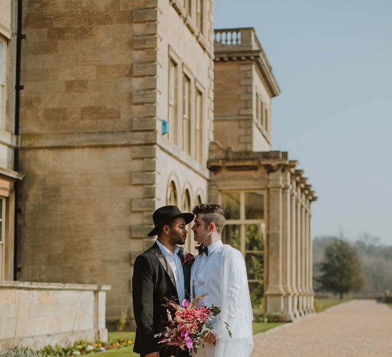 Groom in a two piece black suit and fedora hat standing on the steps at Prestwold Hall with his partner in a white three-piece groom suit, white boots and sheer cape 