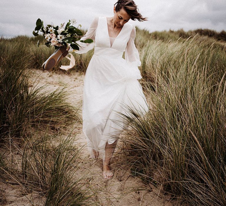 Bride holds her bouquet and wedding shoes as she walks in sand towards the beach in tulle Sassi Holford wedding dress