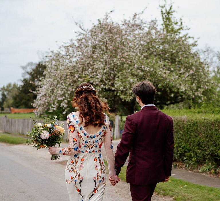 Groom in burgundy suit walks holding hands with bride in colourful wedding dress and bouquet 